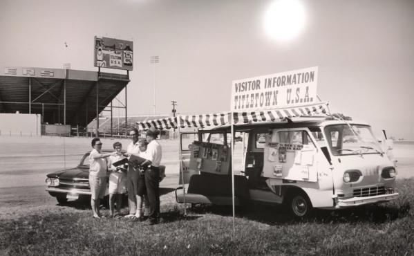 Image of people standing out side of Lambeau Field in the 1960s handing out visitor information from a mobile visitor center.