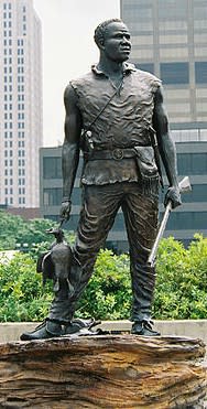 Statue of York, a member of the Lewis and Clark Expedition, standing with a rifle and supplies at his side in an outdoor plaza with city buildings in the background.