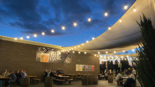 Photo shows beer garden with string lights, a sign that reads "Hella Local" and people sitting at tables drinking craft beer