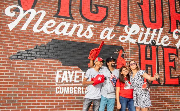 Group of people posing in front of a mural reading “Victory Means a Little More Here” in Fayetteville, Cumberland County, NC