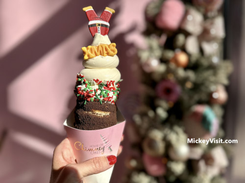 Image of a dessert being held up in front of a pink wall, with a holiday tree in the back right-hand corner of the image. The dessert looks to be an ice cream cone with a decoration that reads 'XMAS' in gold lettering. Red, white, and green sprinkles can be seen under the ice cream swirl. The cone appears to be soft and chocolate flavored.