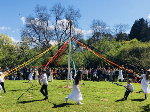 People dance joyfully around a maypole with colourful ribbons on a sunny day. A crowd watches, surrounded by lush greenery and clear skies.