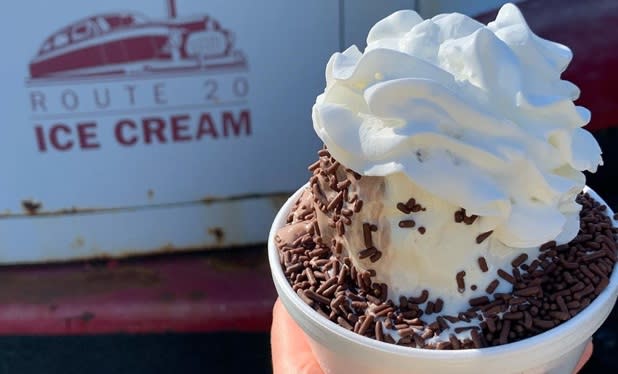 Vanilla ice cream topped with chocolate sprinkles and whipped cream in front of a sign that reads "Route 20 Ice Cream"