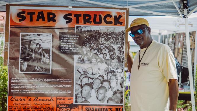 A black man in sun glasses stands beside a banner from Carr's Beach