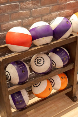 A wooden rack displays various colorful bowling balls, including purple, orange, and white, against a brick wall backdrop. The scene suggests a recreational setting.