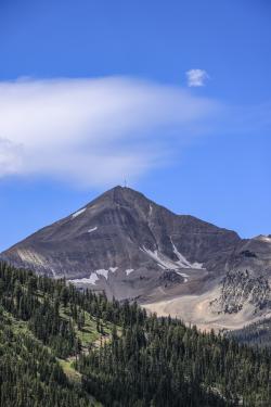 A majestic mountain peak rises against a clear blue sky, surrounded by lush green forests and rocky terrain, inviting exploration and adventure.