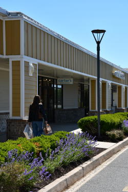 A woman walks in front of a Carter's outlet store at the Bayside Tanger Outlets in Rehoboth Beach, Delaware. There are green bushes and purple flowers in the foreground.