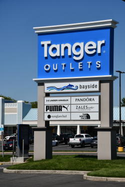 A blue and white sign outside of the Tanger Outlets in Rehoboth Beach lists stores including Pandora, Puma, Zales, Sunglass Hut, and The North Face.