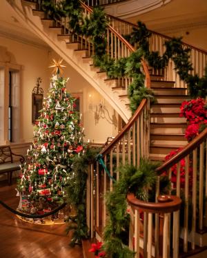 A Christmas tree sits at the bottom of a winding, wooden staircase in the mansion at Winterthur Museum. The staircase railings and steps are decorated with green garland and poinsettias.