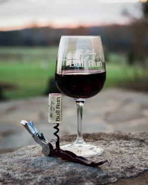 a glass filled with red wine sitting atop historic ruins at the winery at bull run, with a corkscrew holding a popped cork