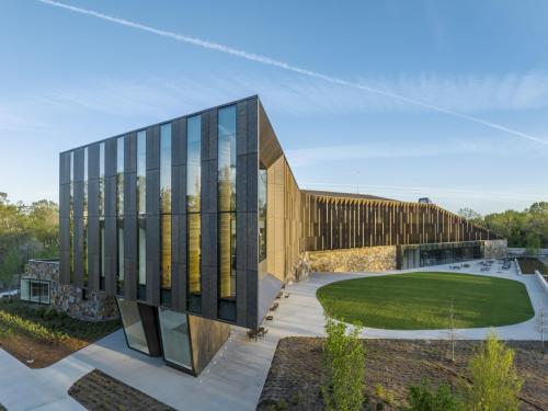 A modern, angular building with dark wood or metal panels and large glass windows, featuring stone accents. An oval green lawn and paved patio with outdoor seating are in the foreground, set against a clear blue sky.