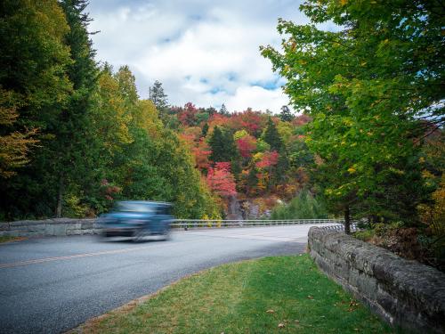 A blue car zooms by on a road with stone walls on each side, with a pop of autumn leaf color in the mountains behind.