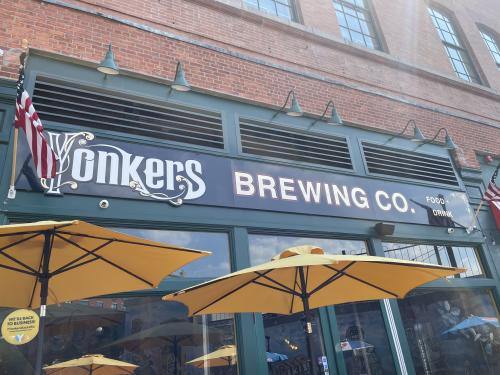 The exterior green sign of Yonkers Brewing Co. is shown. In front of the sign are open yellow umbrellas at outdoor seating tables.