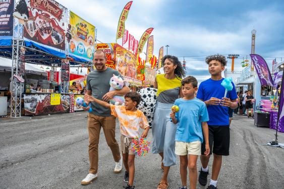 A smiling family with three kids walks happily at the Great New York State Fair, holding cotton candy, toys, and prizes. Bright vendor stalls and banners line the background.