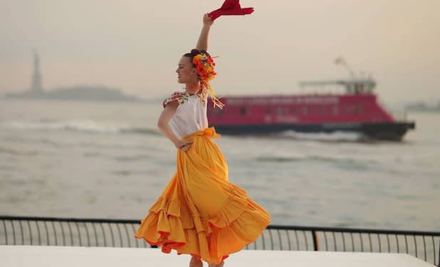 A woman in a flowing yellow skirt and white shirt twirls with a red cloth in her hand in front of the backdrop of a red boat and the Statue of Liberty in the distance