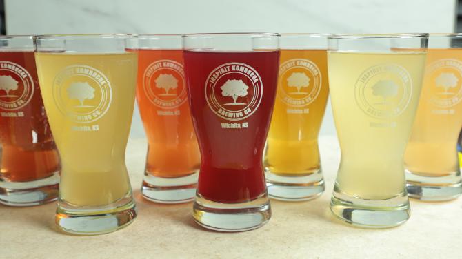 Seven glasses with different colored rinks sit on a table. All have logos that read "Inspirit Kombucha Brewing Co. Wichita KS"