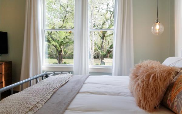 View of a room at Abita Springs Hotel showing a bed with a fluffy pillow and two windows that frame a pair of oak trees outside.