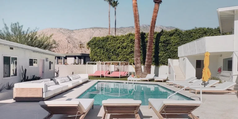 A serene poolside area of a modern house with plush white loungers, a background of mountains, and palm trees under a clear sky. A pink cabana and a hanging yellow towel add pops of color to the scene.