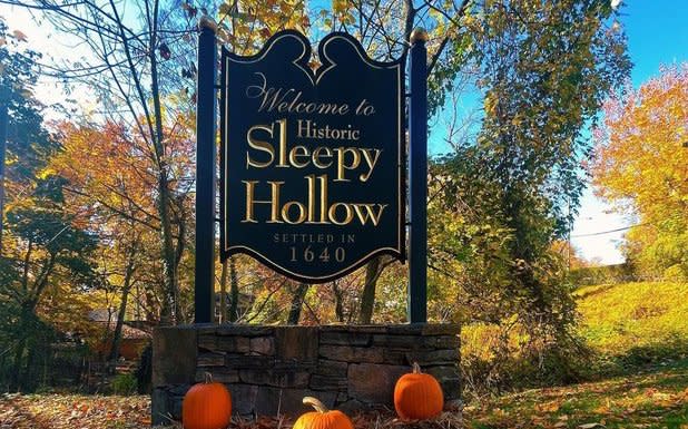 A black sign that reads "Welcome to Historic Sleepy Hollow Settled in 1640" in gold writing. three orange pumpkins sit in front of the sign, backdropped by yellow and orange fall foliage