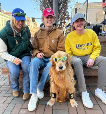 Three people posing with a dressed up and in sunglasses golden retriever at Goldens in Golden CO event 2025