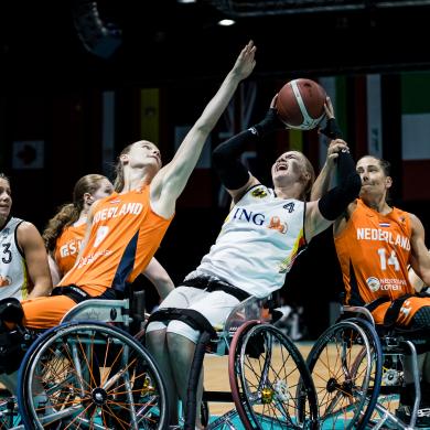 Several athletes in sports wheelchairs compete for a basketball on an indoor court. Two athletes in bright orange jerseys with “NEDERLAND” visible and one in a white jersey with “ING” reach upward toward the ball at the same moment, arms extended. Additional players in white and orange uniforms cluster closely around them. The scene is tightly framed on the action, with visible wheelchair frames, angled wheels, and protective gear. In the background, a row of international flags hangs above the court under dim arena lighting.