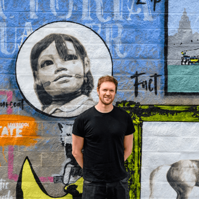 A phot of a man standing infront of a wall with a mural with Liverpool images on.