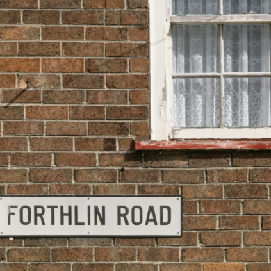 A brick wall on a house with a window to the top right. There is a drain pipe to the left with a sign for Forthlin Road.