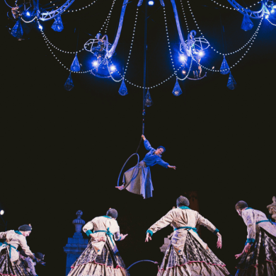 A large chandelier hanging above a group of dancers with people performing ariel dancing from it.