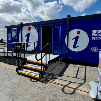 A visitor welcome centre inside a large shipping container style venue. It is painted dark blue and has a ramp leading up to thedoor. There is a large logo of an i on the branding.