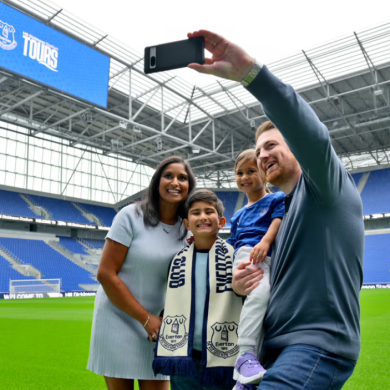 A family of two adults and two children with one adult standing taking a selfie style photo of the family with the Everton pitch behind them.