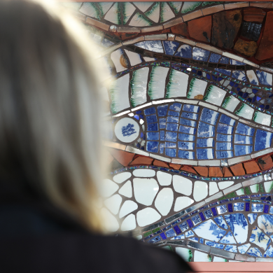 A person viewed from behind looks at a colourful mosaic artwork made of broken tiles and ceramic pieces. The mosaic features flowing, wave‑like patterns in blue, green, white, and earthy tones, arranged in curved bands against a brick wall.