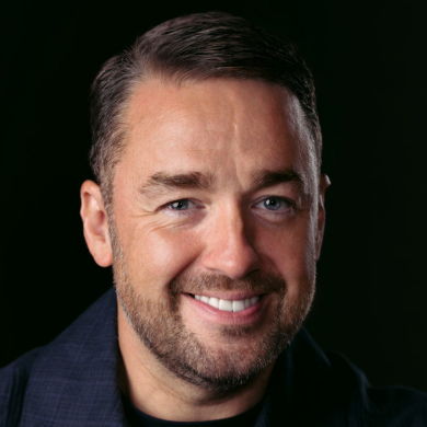 A man pictured smiling brightly wearing a navy jacket with a black background.