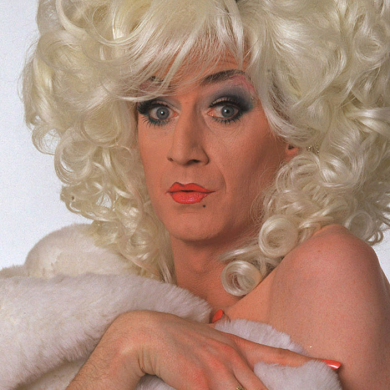 A person with a large, voluminous curly blonde wig poses wrapped in a white fur-like garment. The person has dramatic eye makeup, bright lipstick, and long painted nails, looking directly toward the camera against a plain background.