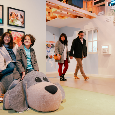 A girl and a boy sit smiling on a large, soft, dog-shaped seat in a brightly lit, family-friendly exhibition space. Behind them, photos of animals hang on the wall. In the background, two adults walk through the room, observing the displays.