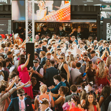 A large, crowded indoor event space filled with people dressed in formal and semi-formal attire, gathered around bars and food stalls. Overhead screens and signage read “Eat. Drink. Play.” as attendees mingle, raise drinks, and socialise in a lively atmosphere.