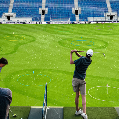 Indoor stadium transformed into a golf driving range, with a person swinging a golf club from a raised platform toward circular targets marked on the green field below, while others watch from nearby seating.