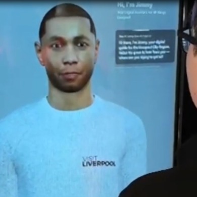 Person standing in front of a large digital screen displaying a lifelike virtual avatar wearing a light blue top with “Visit Liverpool” text, suggesting an interactive or AI‑powered information display in an indoor public setting.