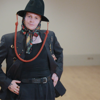 A person standing in an empty indoor room with a wooden floor and plain walls, wearing a dark blazer with decorative pins, a black hat with red cords, a patterned scarf tied under the chin, and black trousers, posing with hands on hips.