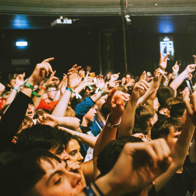 A densely packed indoor crowd with many arms raised above heads, people wearing wristbands and holding phones, photographed from within the audience under warm stage lighting at a live event or concert.
