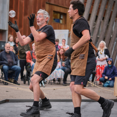Outdoor performance in a public space showing two performers standing on a small platform, both wearing brown work-style aprons, black clothing, gloves, and boots. One performer tosses a clear glass object into the air while the other steps forward nearby. A seated audience watches from behind, with industrial-style buildings, wooden structures, and equipment visible in the background.