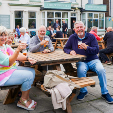 Outdoor pub terrace scene with several people seated at wooden picnic tables, holding drinks and socialising outside a building labelled ‘The Windmill Public House,’ with bunting decorations and other guests gathered in the background.