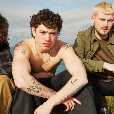 Group portrait of four people sitting outdoors on grass near a body of water. The group faces the camera in a close formation, wearing casual clothing including jackets, trousers, and trainers. One person at the centre is shirtless with visible arm tattoos and a chain necklace. The background shows open sky and a coastal landscape under natural daylight.
