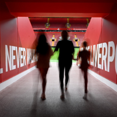 Three people walking down the path to the Anfield pitch with white writing saying 'You'll never walk alone' on a red background.