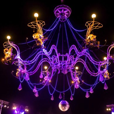 A large chandelier lit up in purple lights with performers on it as part of an act.
