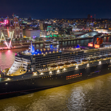A large cruise liner ship on the River Mersey in Liverpool. It is night time and the skyline of the city is behind it. There are fireworks coming from the Pier.