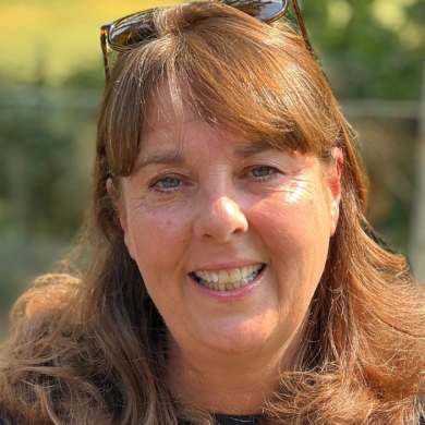 A woman sitting smiling looking at the camera, with brown hair and black sunglasses on her head.