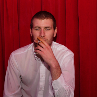 A man smoking a cigarette, wearing a white shirt with a red backdrop behind him.