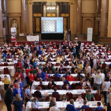 A row of tables all set out in St George's Hall with people sitting at the tables playing chess. There is a large screen at the centre of the room with a chess board featured on it.
