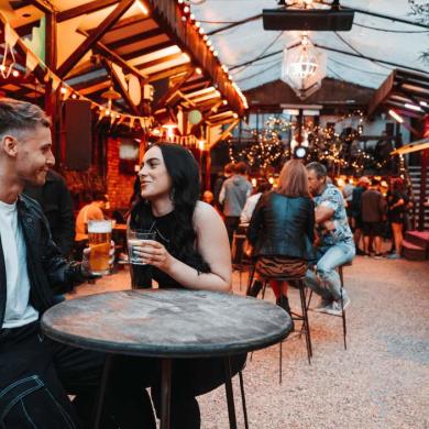 Indoor food and drink venue with warm ambient lighting, wooden market-style stalls and decorative string lights. In the foreground, two visitors sit at a round table holding drinks, while other guests gather and socialise throughout the space. The covered courtyard features a mix of seating areas, plants and illuminated vendor units, creating a lively and inviting atmosphere for an evening out.