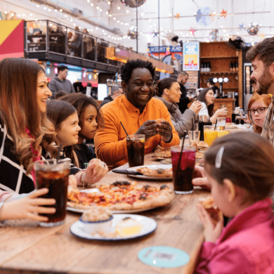 A family are sitting at bench tables in a food market. There are different coloured food vendor stalls in the food market.